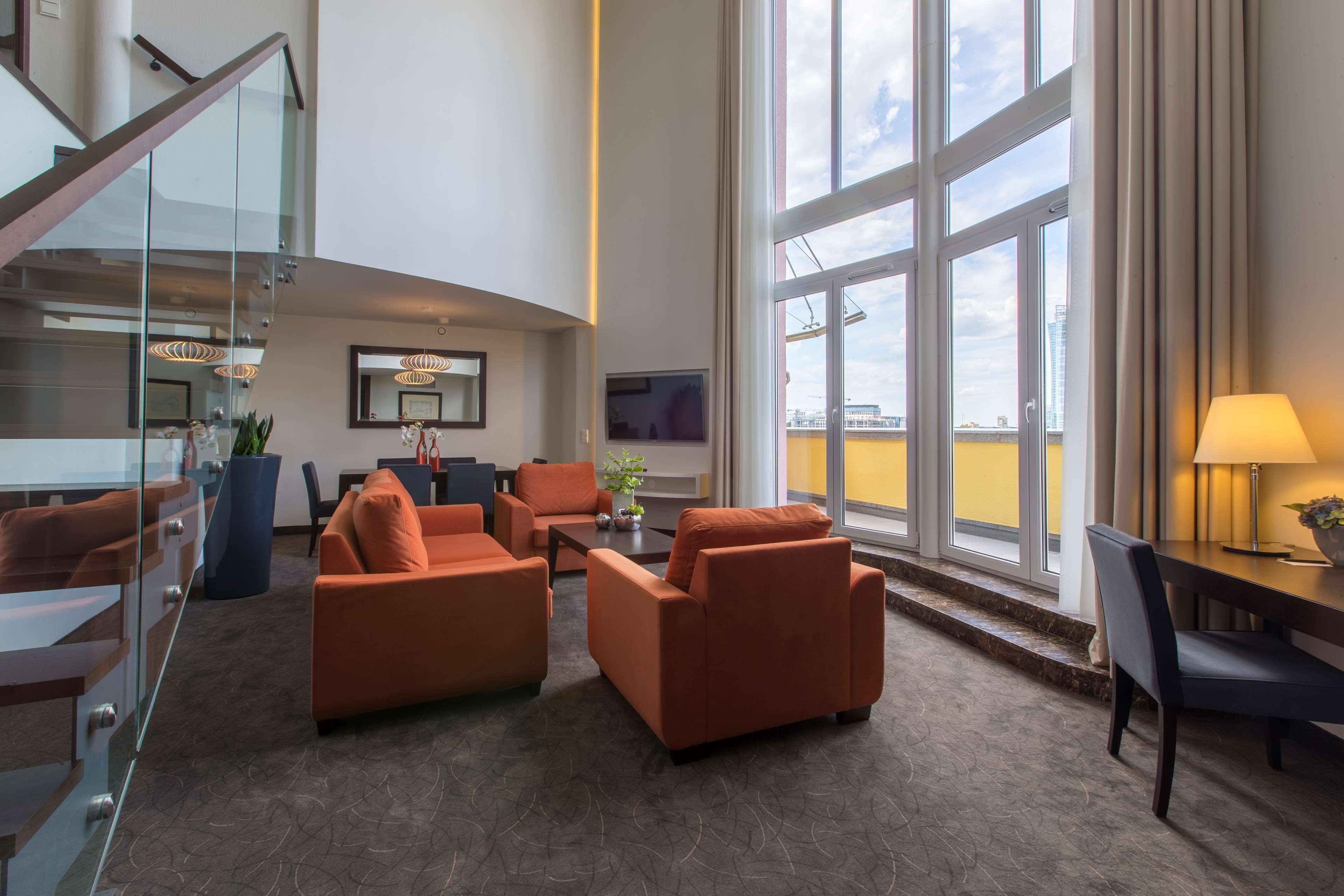 Radisson Blu Sobieski Warsaw Exterior photo The photo shows an elegant and spacious living area. There are four orange chairs arranged around a coffee table, creating a cozy seating space. A large window with multiple panes allows natural light to flood the room, and there are tall curtains th