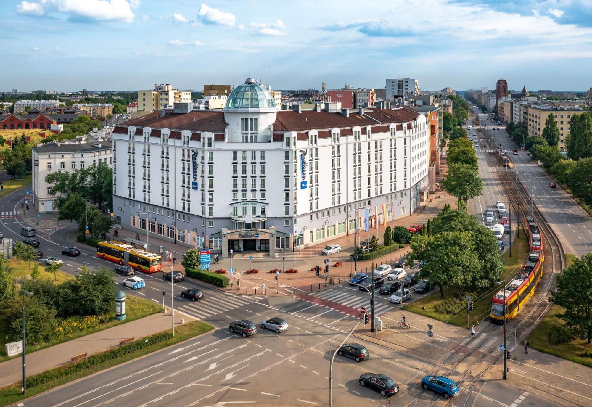 Radisson Blu Sobieski Warsaw Exterior photo The photo shows a large, modern building with a distinctive architectural design, featuring a rounded dome on top. It's situated at a crossroads, surrounded by greenery and several roads. In front of the building, there are a few trams visible, indic